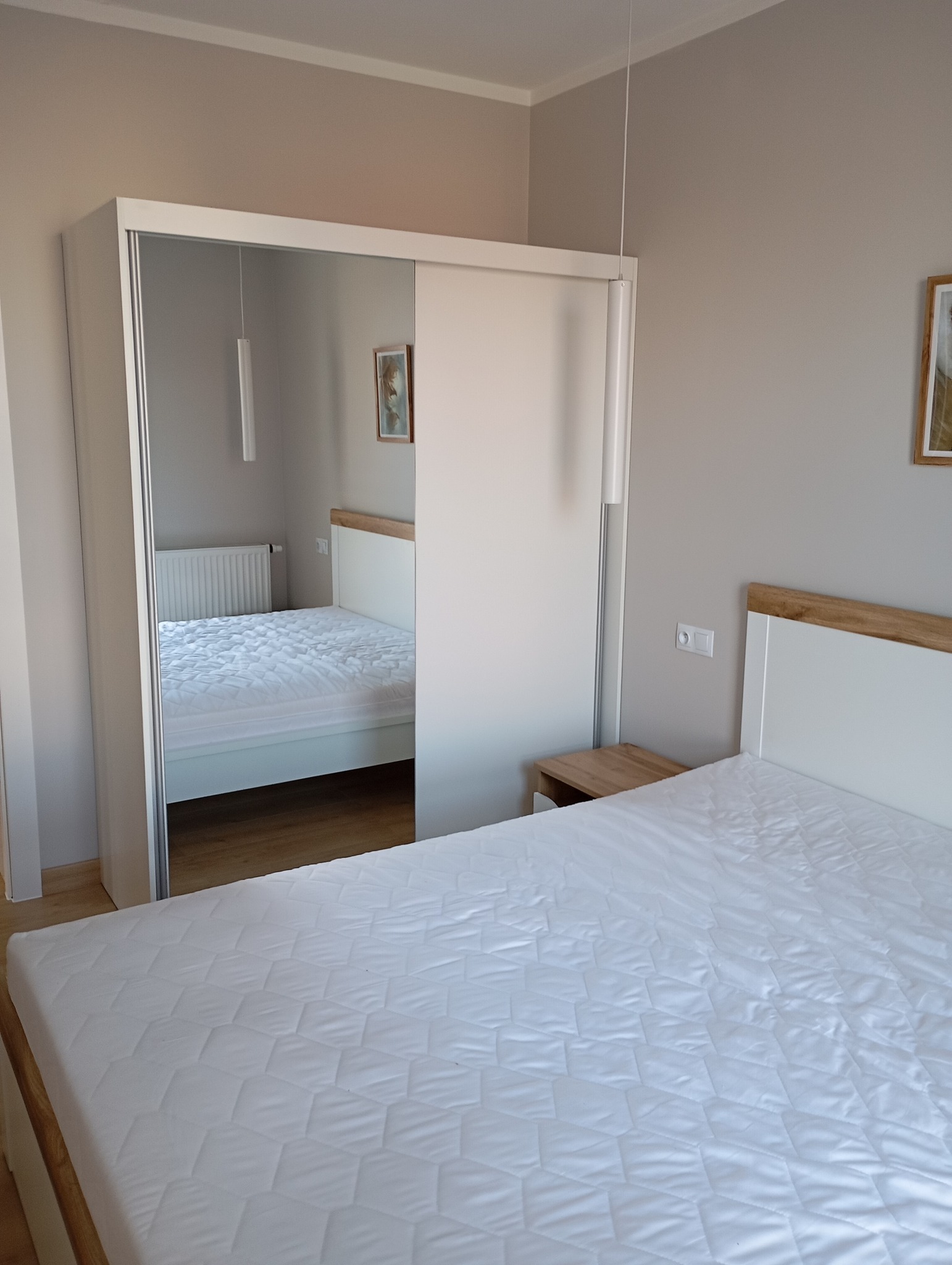 Nowoczesna sypialnia z białym łóżkiem i szafą przesuwną z lustrem, odbijającym fragment pokoju. Jasne ściany i drewniane akcenty dodają przytulności.