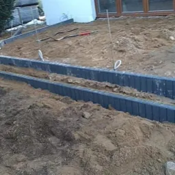 Układanie obrzeży z kostki brukowej w ziemi, widoczna łopata z pomarańczowym elementem odblaskowym, ziemia, piasek i pracownik w odzieży roboczej.