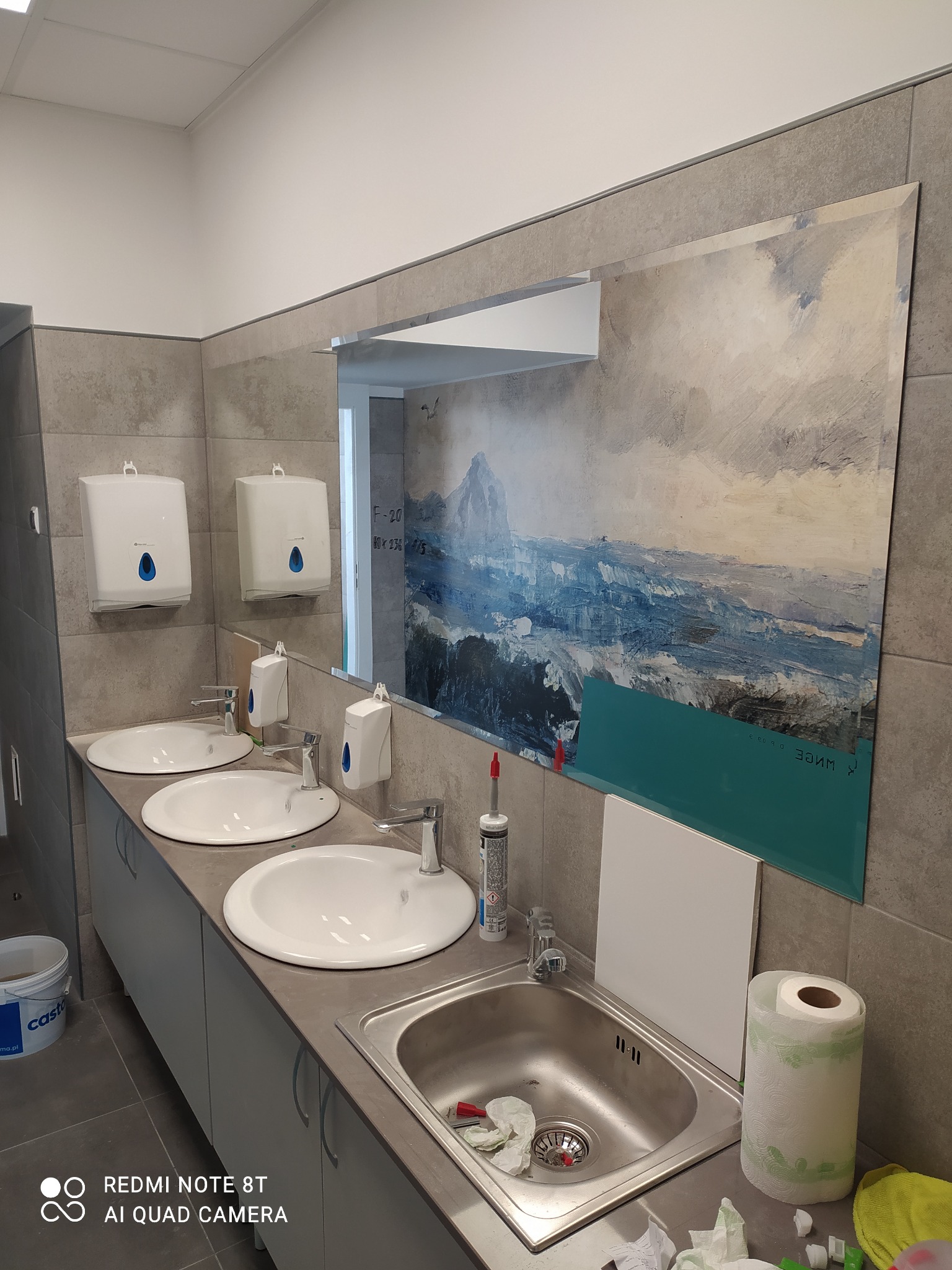 Publiczna łazienka z trzema umywalkami i dużym lustrem z motywem pejzażu górskiego, częściowo zakrytym turkusową folią ochronną, widoczny silikon i rolka ręcznika papierowego na blacie.