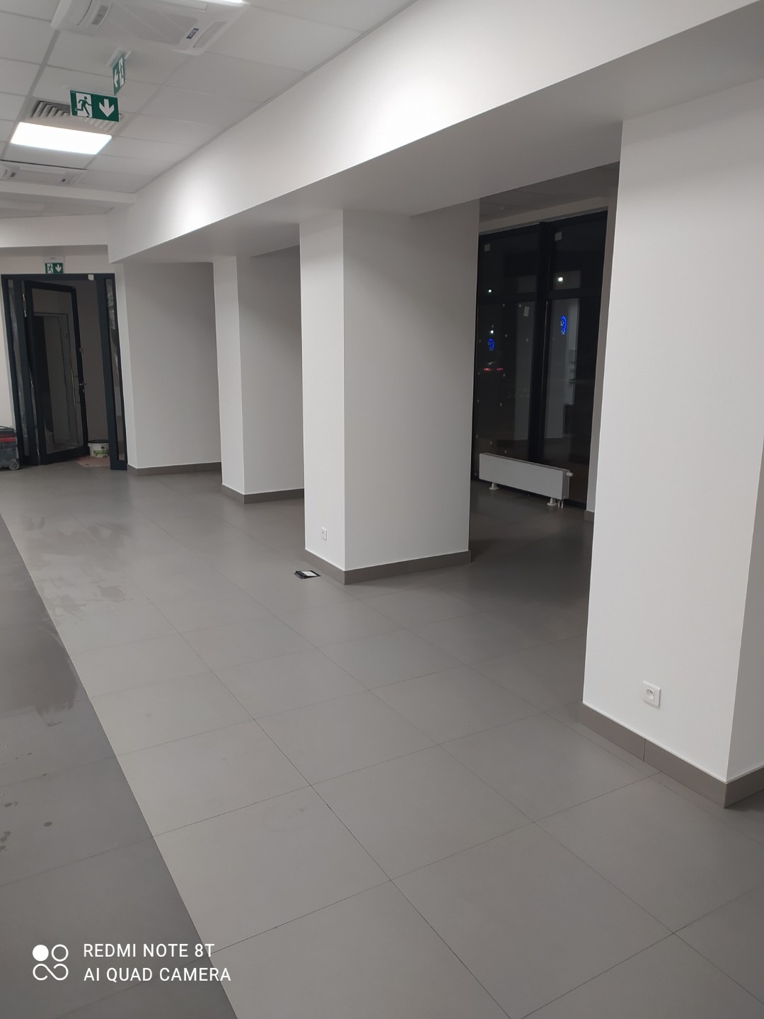 Wnętrze biurowe po malowaniu, widoczne białe ściany, szara podłoga z płytek, kolumny i okna z widokiem na nocne miasto. Na suficie widoczne znaki ewakuacyjne.