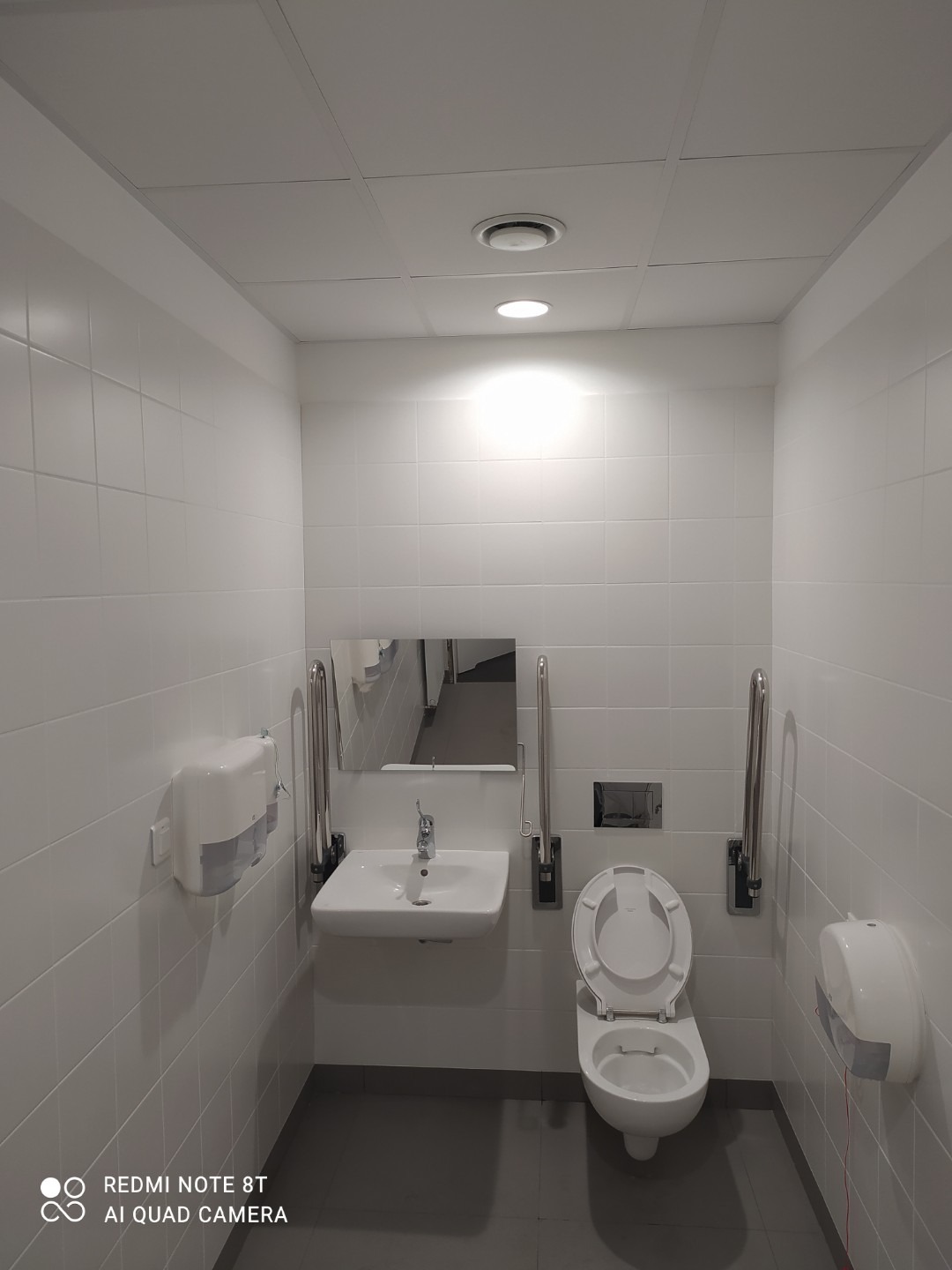 Biała łazienka publiczna z toaletą, umywalką i uchwytami dla niepełnosprawnych, wykończona białymi płytkami.