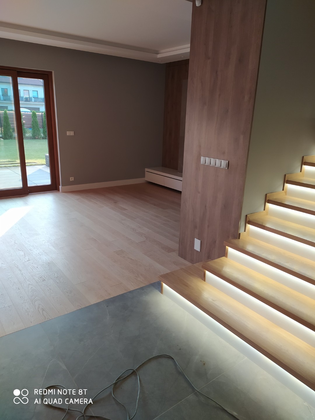 Nowoczesne wnętrze z drewnianą podłogą, szarymi ścianami i schodami z podświetleniem LED, widok na ogród przez duże okno.