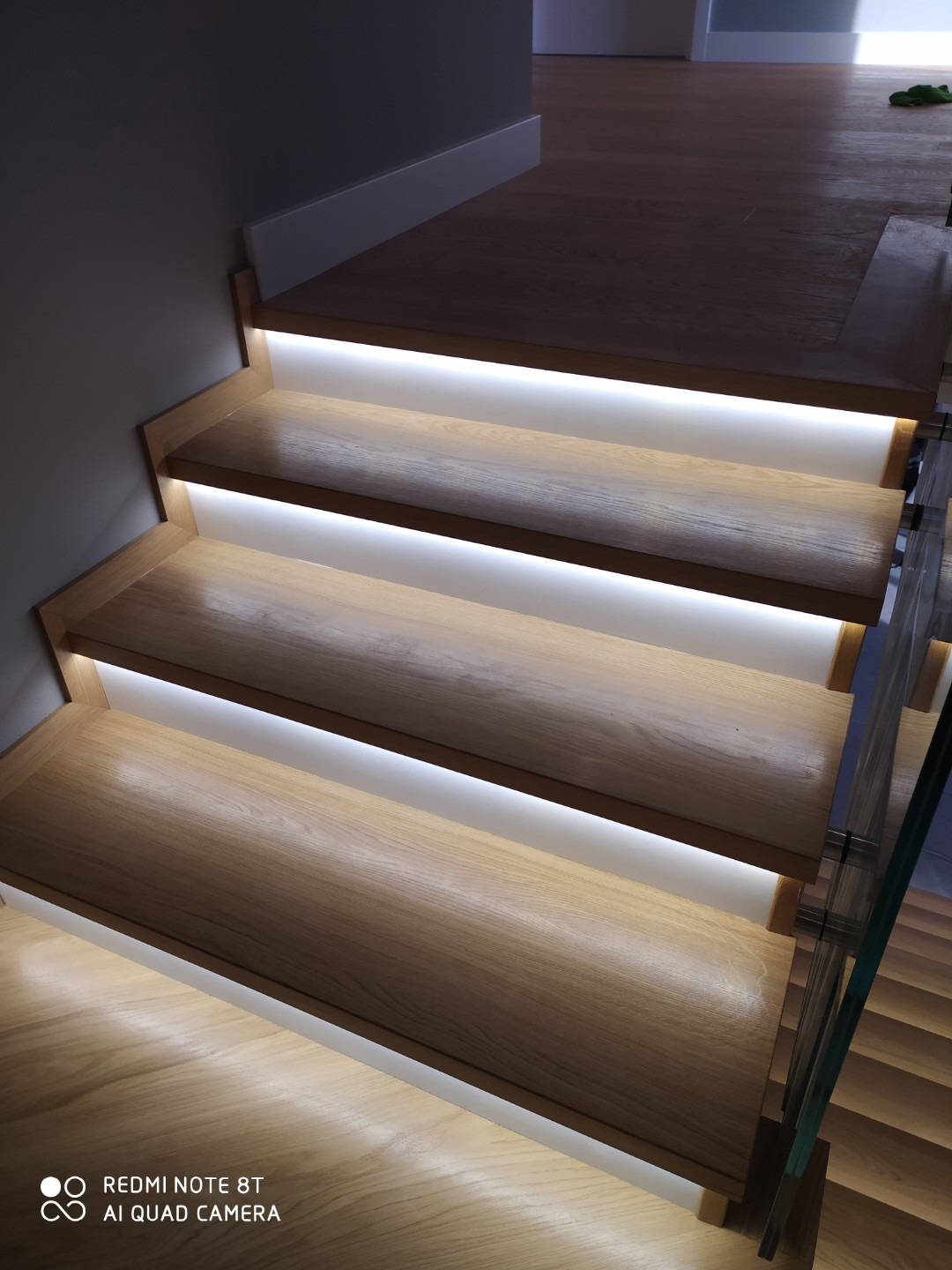 Drewniane schody z podświetleniem LED, szklana balustrada, widok z dołu, nowoczesny design.