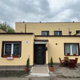 Budynek ośrodka szkoleniowego z tablicą informującą o szkoleniach operatorów maszyn budowlanych i drogowych, widoczne okno z kwiatami, drzwi wejściowe z szyldem K-MILA i stolik z krzesłami...