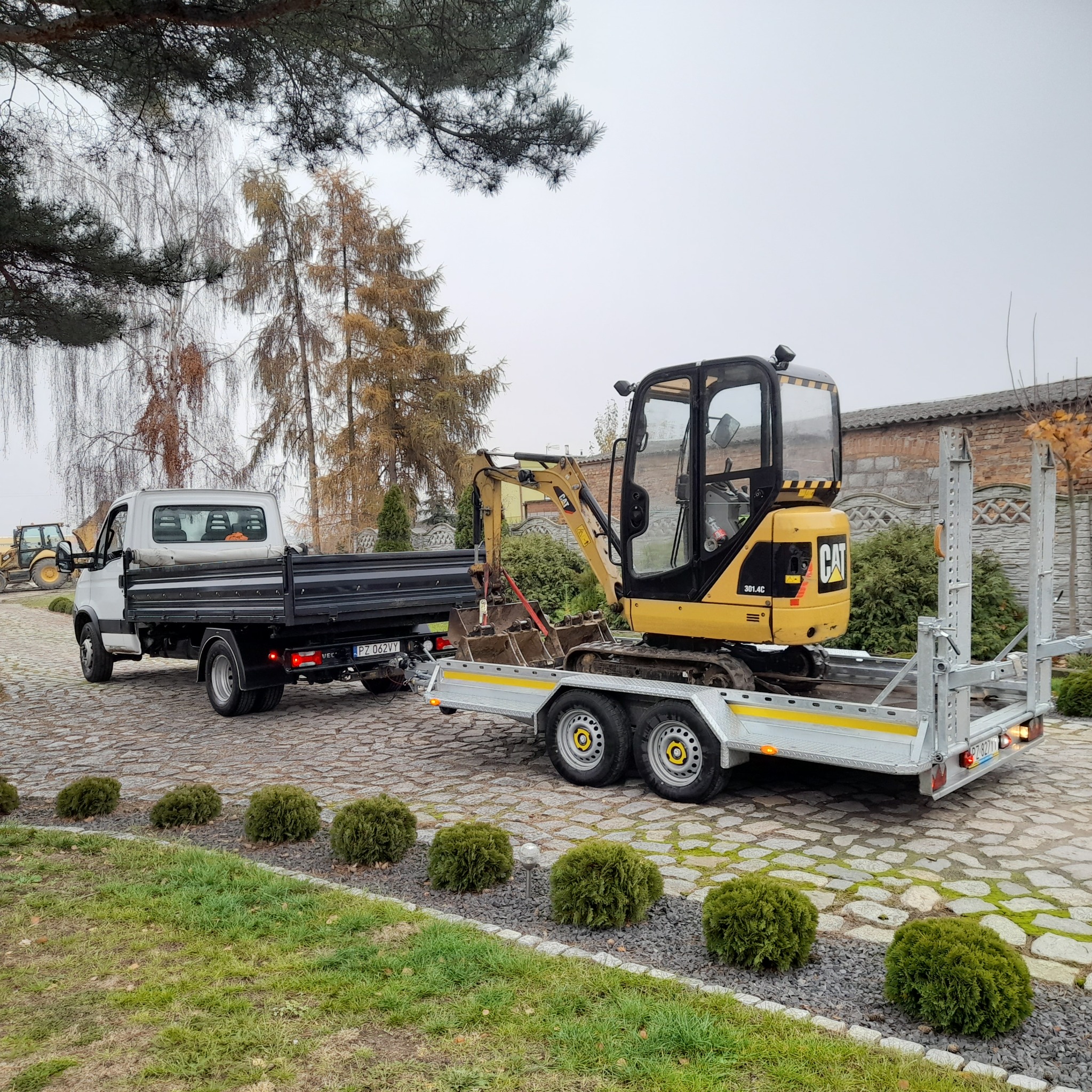 Mała żółta koparka CAT 301.4C przewożona na przyczepie przez białą ciężarówkę na tle brukowanej drogi i zielonych krzewów.