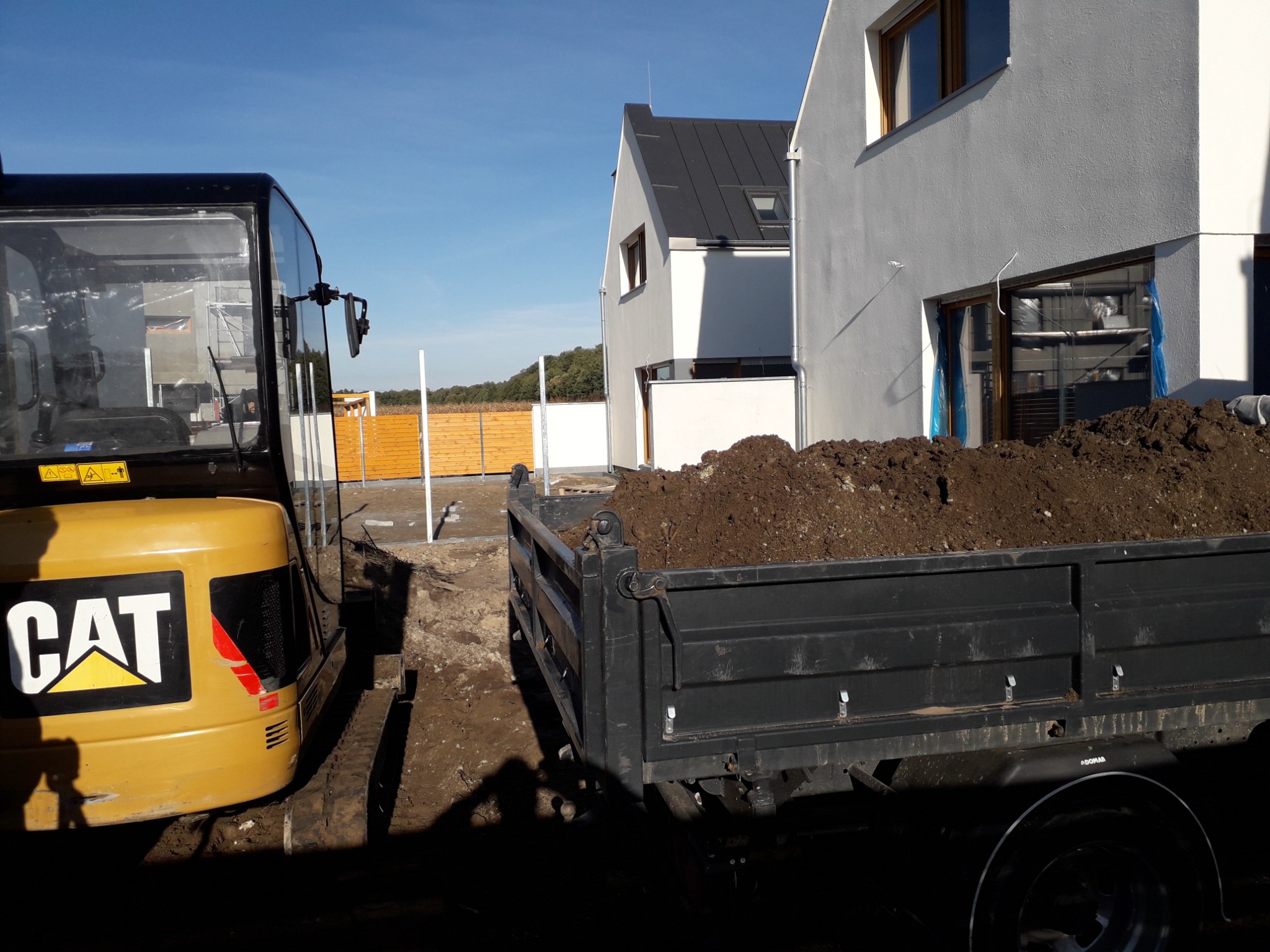 Żółta minikoparka CAT i przyczepa wypełniona ziemią na placu budowy przed nowoczesnym domem z szarą elewacją i drewnianym ogrodzeniem w tle.