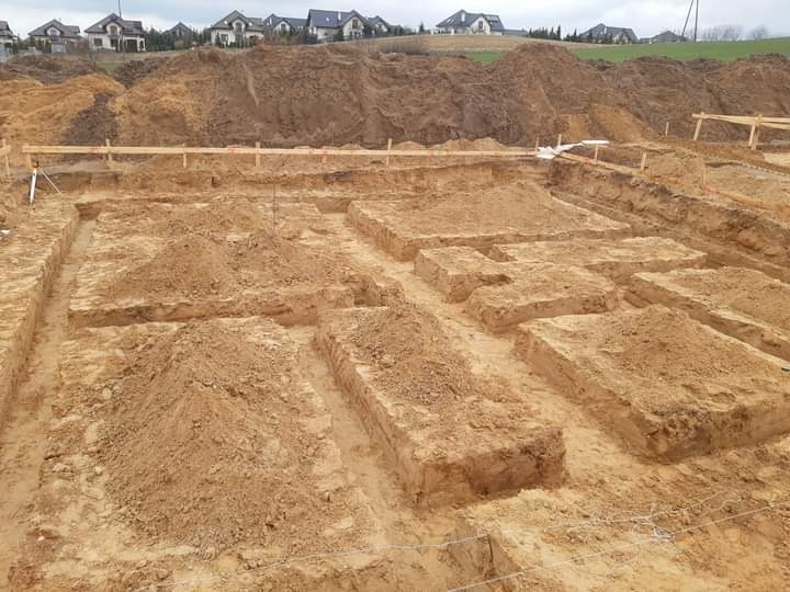 Przygotowany wykop pod fundamenty domu jednorodzinnego, widoczne drewniane szalunki i pryzmy ziemi wewnątrz wykopu, w tle zabudowa mieszkalna.