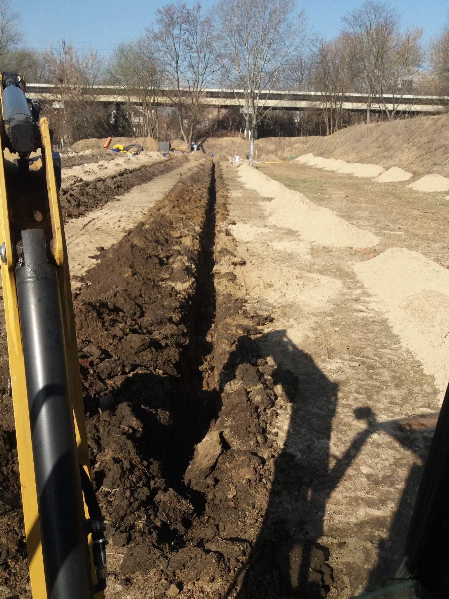 Długi, wąski wykop w ziemi, widoczny z kabiny koparki, z hałdami piasku po bokach i wiaduktem w tle.