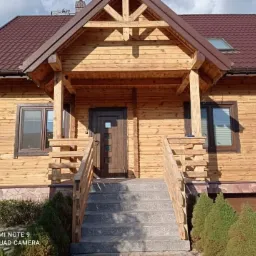 Drewniany dom z brązowym dachem, widok z przodu, schody wejściowe z szarego kamienia, elewacja pokryta lakierem bezbarwnym, widoczne słoje drewna.