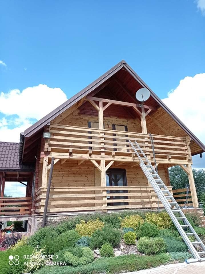 Nowo wybudowany, surowy drewniany dom z widoczną drabiną opartą o balkon i anteną satelitarną na dachu, otoczony zielenią.