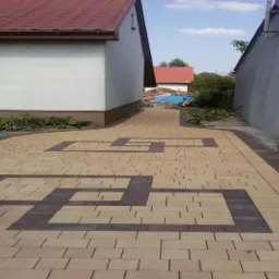 Ułożona beżowo-brązowa kostka brukowa tworząca wzór geometryczny przed domem z czerwonym dachem, widok z perspektywy chodnika.