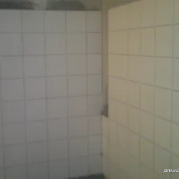 Ściana wyłożona białymi, kwadratowymi płytkami w łazience, widoczne fugi w kolorze szarym, na górze ściany widoczne ślady po remoncie.