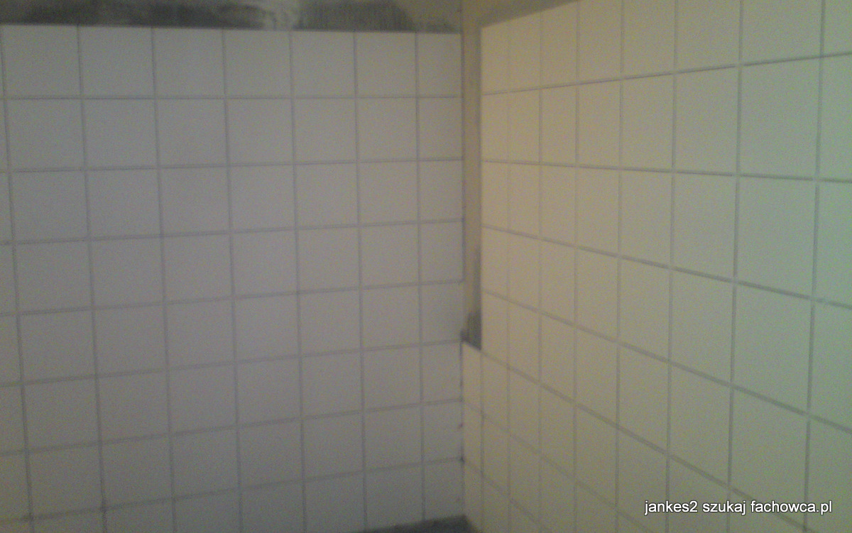 Ściana wyłożona białymi, kwadratowymi płytkami w łazience, widoczne fugi w kolorze szarym, na górze ściany widoczne ślady po remoncie.