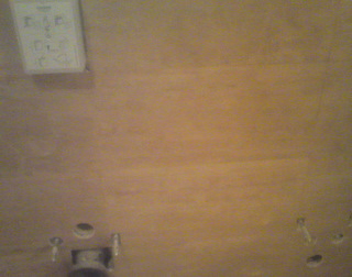 Niejasne ujęcie fragmentu podłogi z widocznymi otworami i elementami montażowymi, z notatnikiem przy krawędzi sufitu. Słabe oświetlenie i niska jakość obrazu.