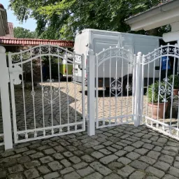 Otwarta, biała, metalowa brama wjazdowa z dekoracyjnymi elementami na posesji wyłożonej kostką brukową, w tle zaparkowany biały bus i osoby w niebieskich koszulkach.