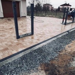 LeticoGroup sp z o.o - Ułożona beżowa kostka brukowa z czarnym obramowaniem, widoczne przygotowania do montażu ogrodzenia, obok leży rozsypany żwir i stoi mieszarka do betonu, w tle dom jednorodzinny i częściowo widoczny...