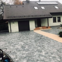 LeticoGroup sp z o.o - Ułożona szara kostka brukowa na podjeździe przed domem z brązową bramą garażową i panelami słonecznymi na dachu, widoczny fragment taczki i pracownik w tle.