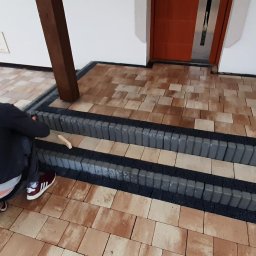 LeticoGroup sp z o.o - Pracownik w trakcie układania stopni z szarej kostki brukowej na tarasie przed wejściem do budynku, widoczne fragmenty ułożonej już kostki w odcieniach beżu i brązu.