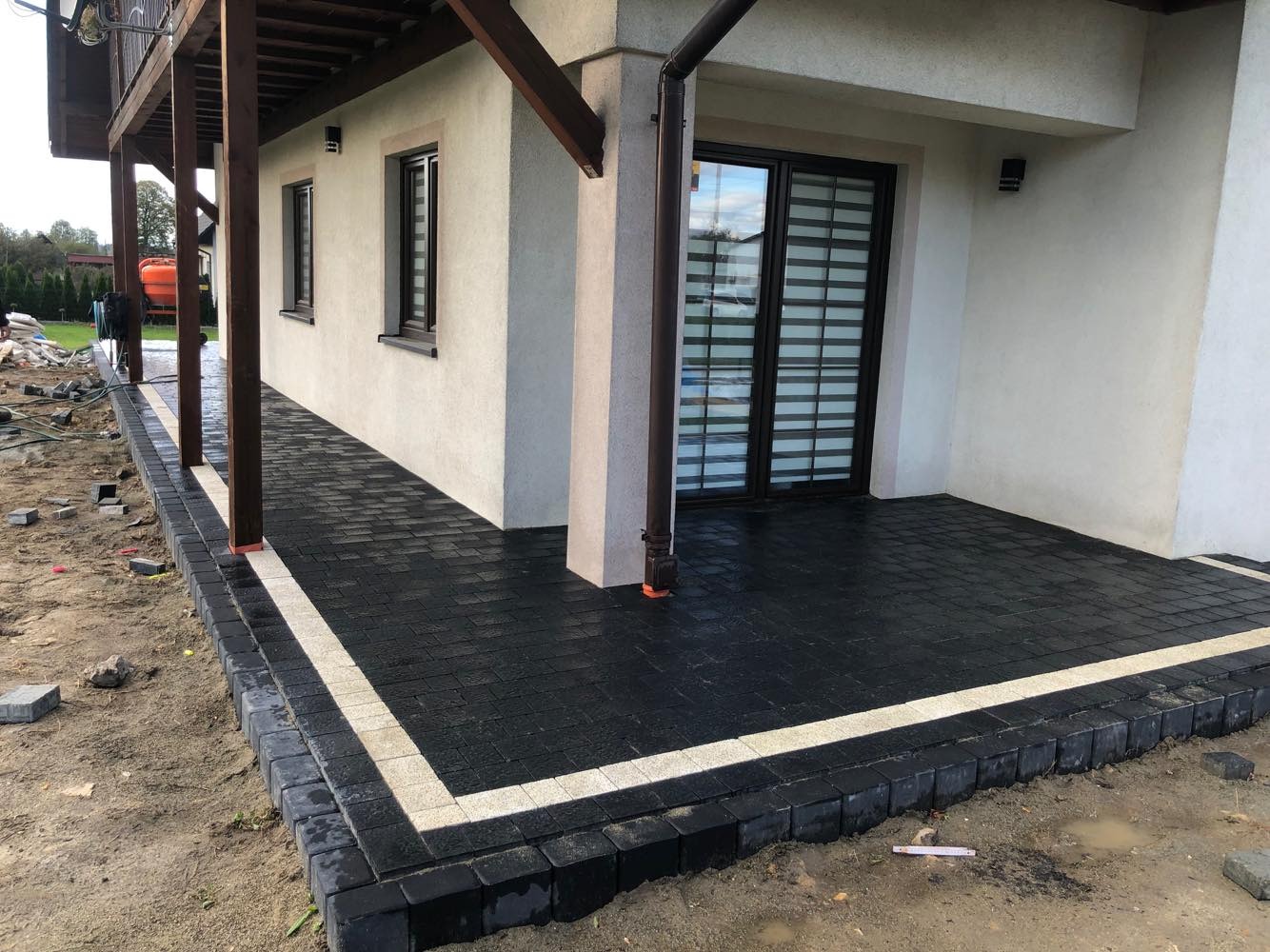 Nowo ułożona, czarna kostka brukowa z jasnym pasem obramowania na tarasie domu jednorodzinnego, widoczne niedokończone prace przy krawężniku i elementy konstrukcji drewnianej.