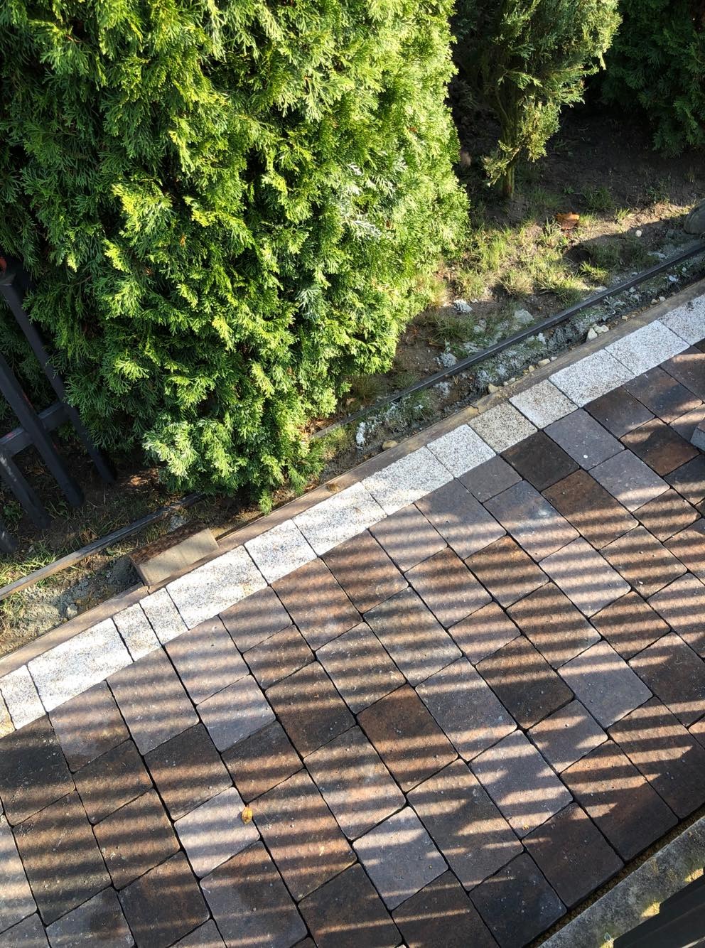 Ułożona kostka brukowa w odcieniach brązu i szarości, obramowana jasnymi kostkami granitowymi, z cieniem rzucanym przez ogrodzenie i zielenią w tle.