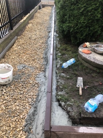 Wykonywanie drenażu liniowego: koryto z brązowego tworzywa sztucznego obsypane drobnym żwirem, widoczna szpachelka, butelka wody i wiadro z zaprawą cementową.