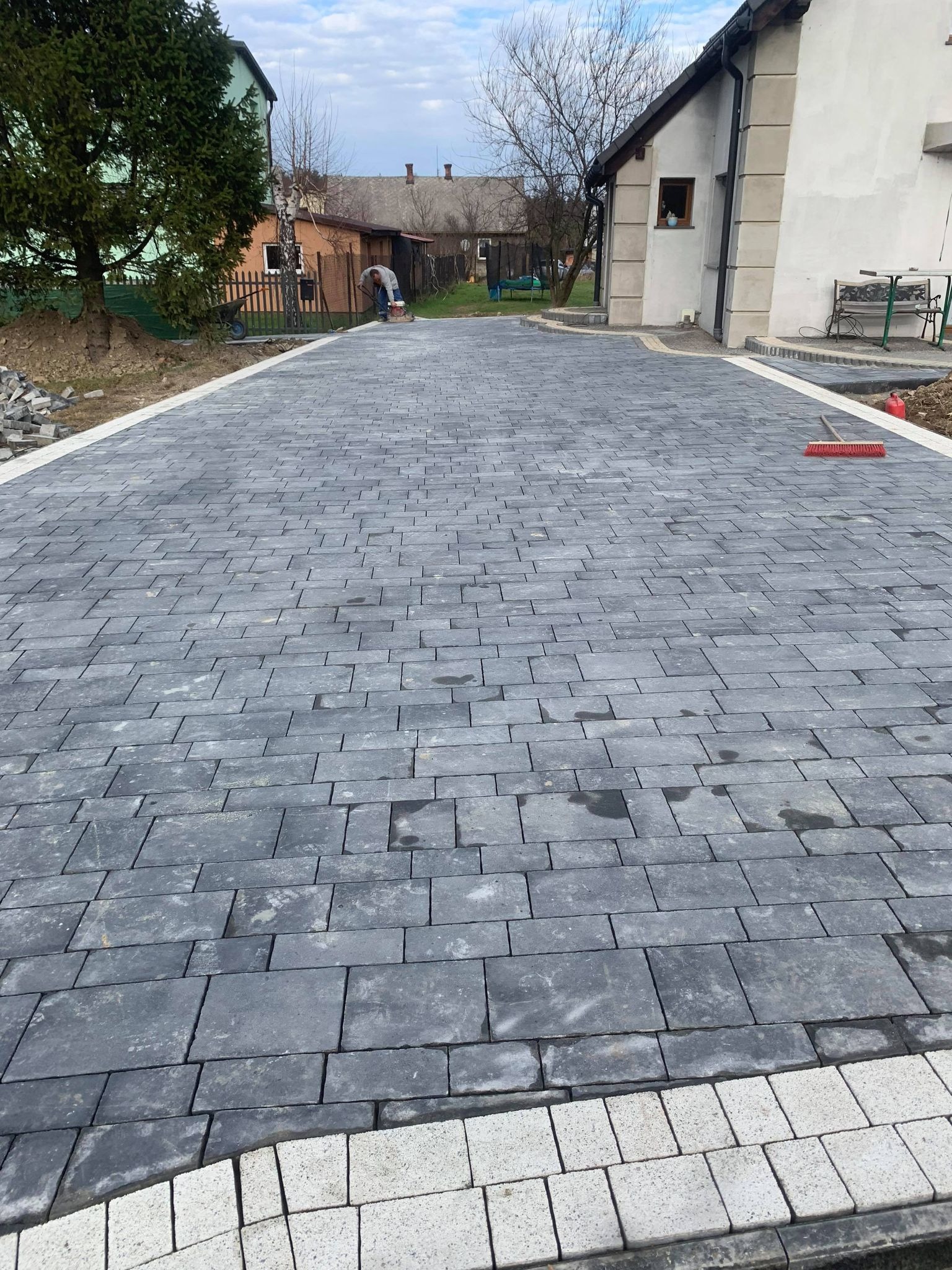 Ułożona szara kostka brukowa na podjeździe domu jednorodzinnego, widoczny pracownik zagęszczający nawierzchnię oraz czerwona zmiotka.