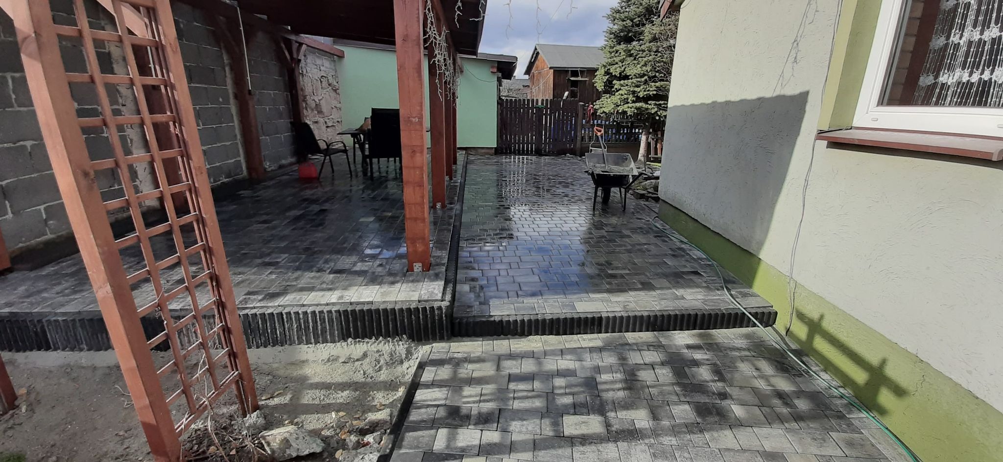 Świeżo ułożona, mokra, szara kostka brukowa na posesji z drewnianą altaną, widoczna taczka z narzędziami i fragment ogrodu.