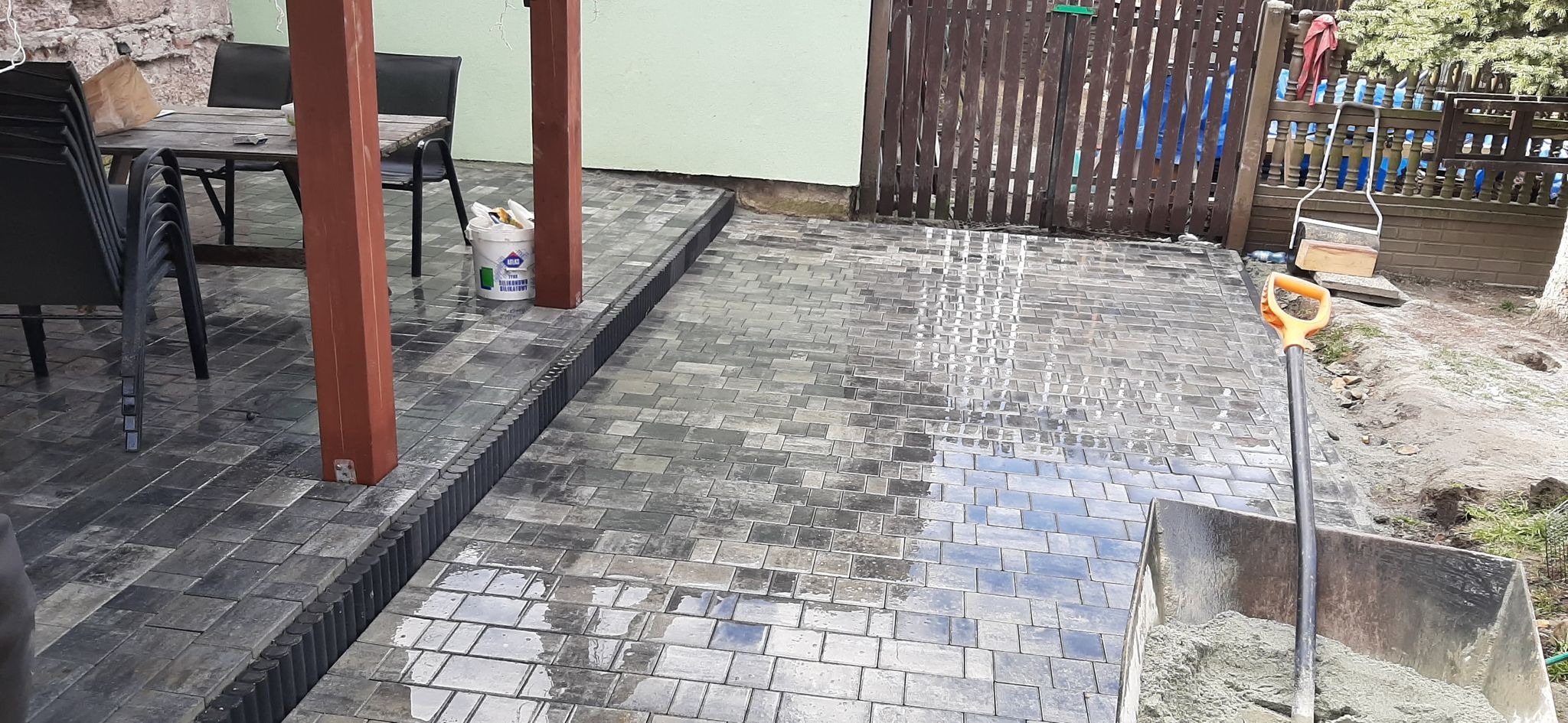 Świeżo ułożona, mokra kostka brukowa w odcieniach szarości, z widocznym korytkiem odwadniającym, opartym o drewniane słupy werandy. W tle widoczny wózek z zaprawą i łopata, oraz narzędzia brukarza.