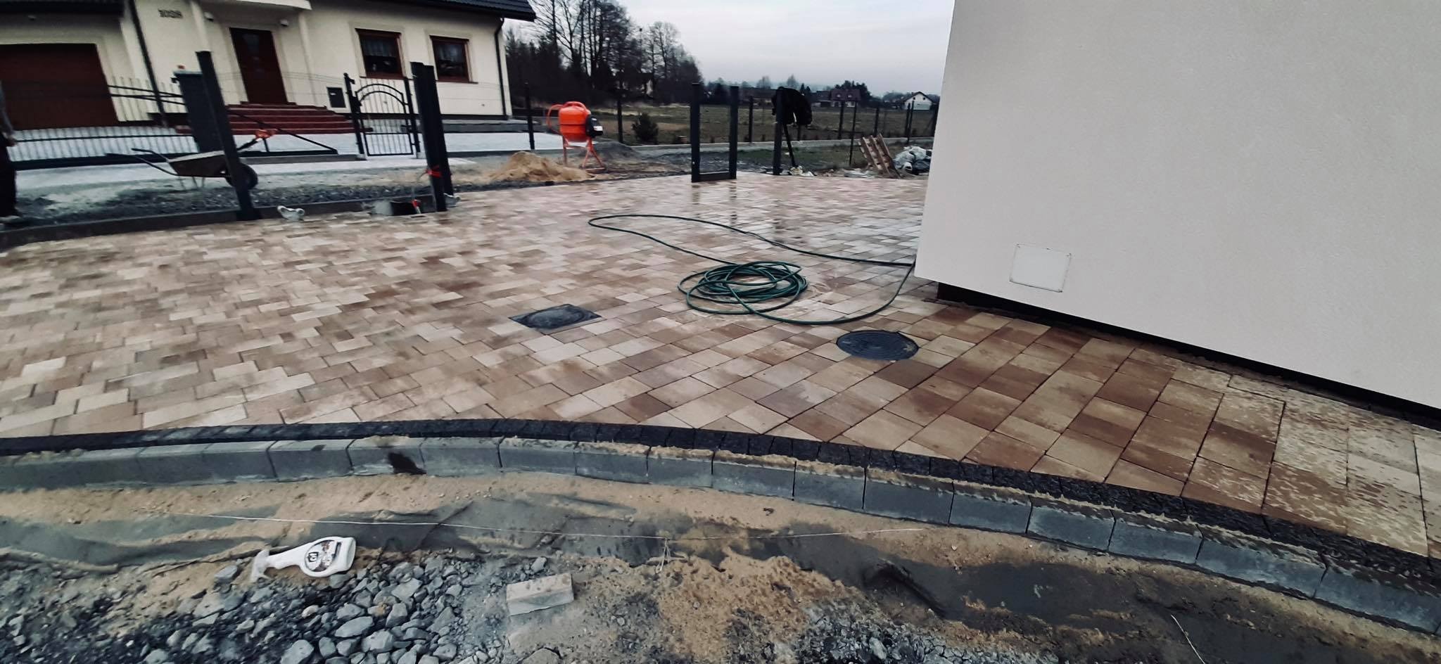 Ułożona kostka brukowa w odcieniach beżu i brązu, z widocznym obrzeżem i zielonym wężem ogrodowym na powierzchni, w tle dom jednorodzinny i betoniarka.