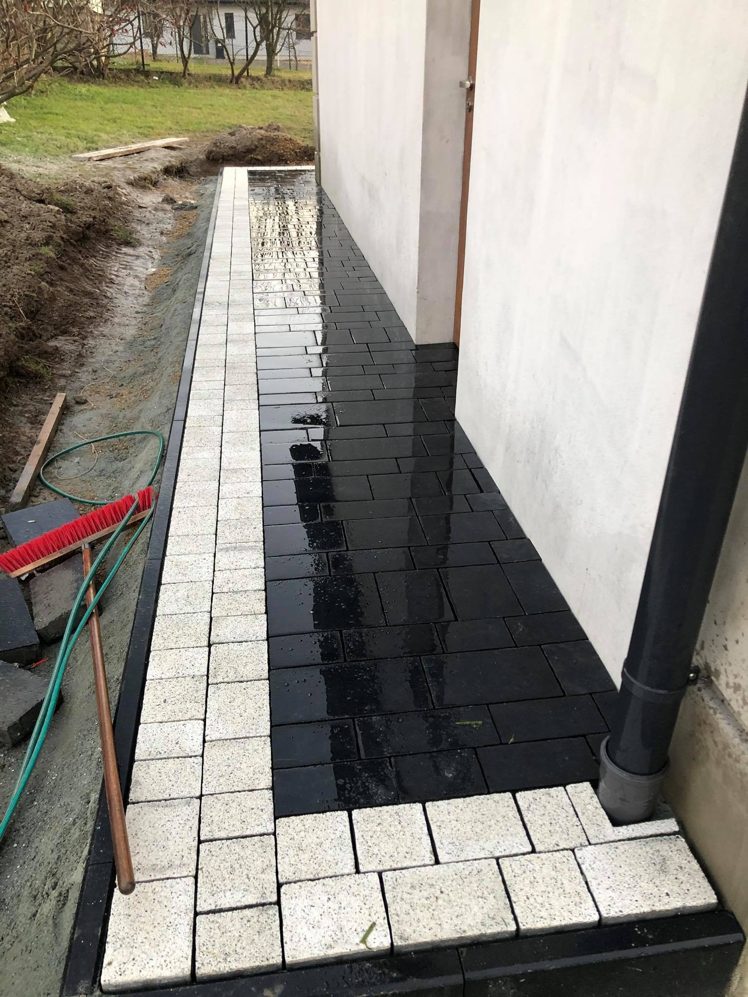 Świeżo ułożona, mokra kostka brukowa w kolorze czarnym i jasnoszarym, tworząca ścieżkę wzdłuż ściany budynku, z widoczną szczotką i wężem ogrodowym w tle.
