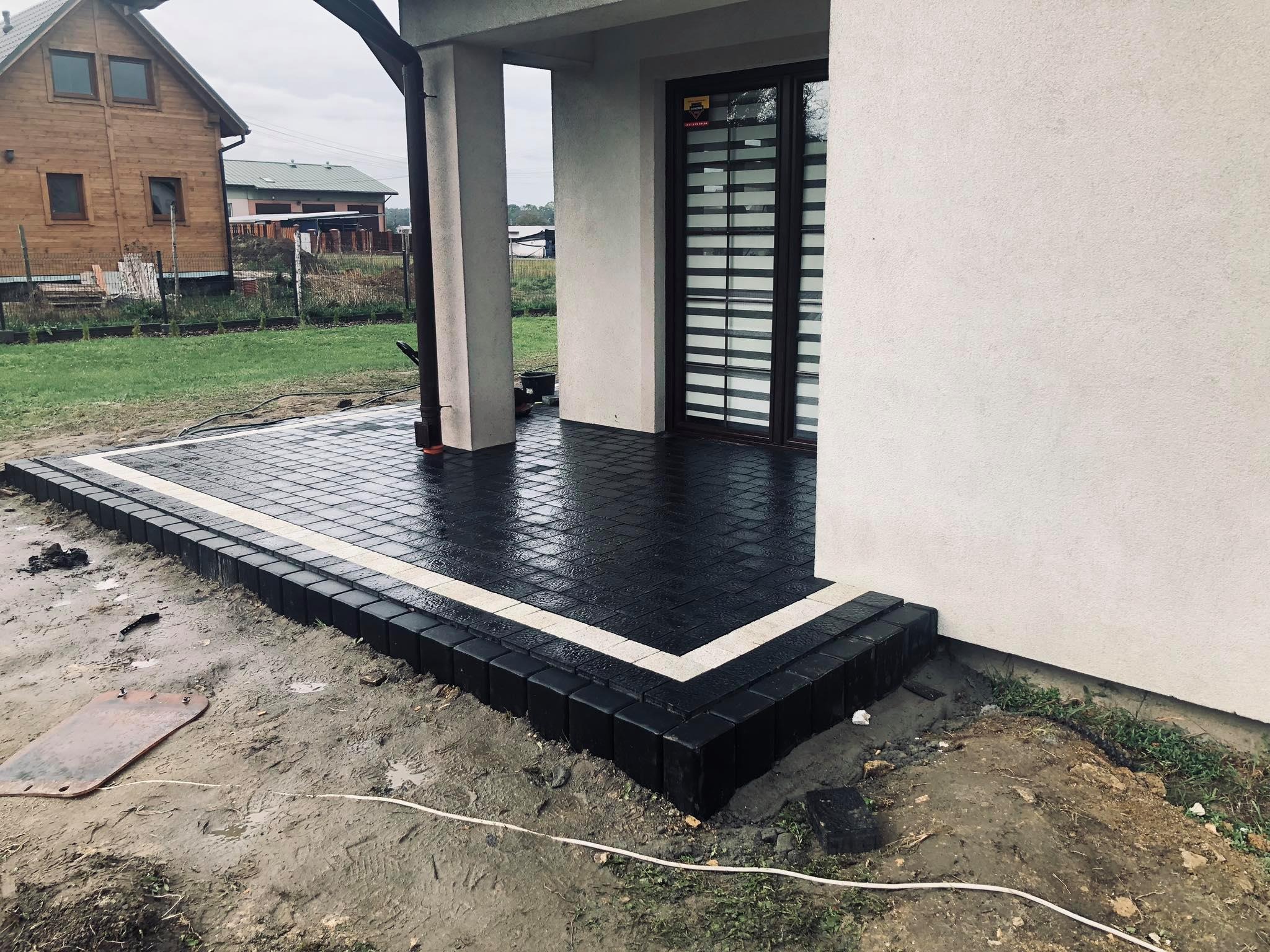 Wykończony taras z ciemnej kostki brukowej z jasną obwódką, przylegający do jasnego budynku z brązowymi oknami, widoczny fragment trawnika i dom w tle.