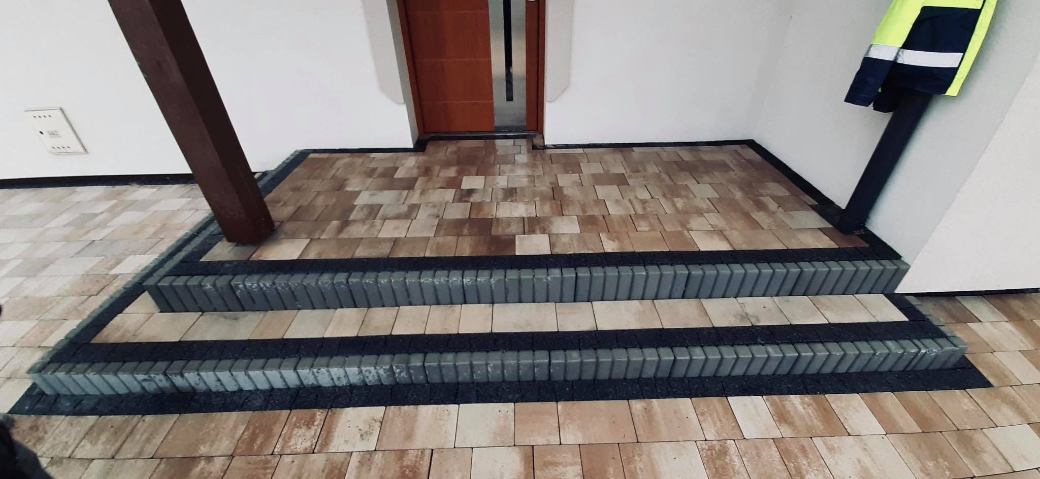 Wykończone schody wejściowe z kostki brukowej w odcieniach beżu i szarości, z widocznym drewnianym słupem wspierającym zadaszenie i elementami odblaskowej odzieży roboczej w tle.