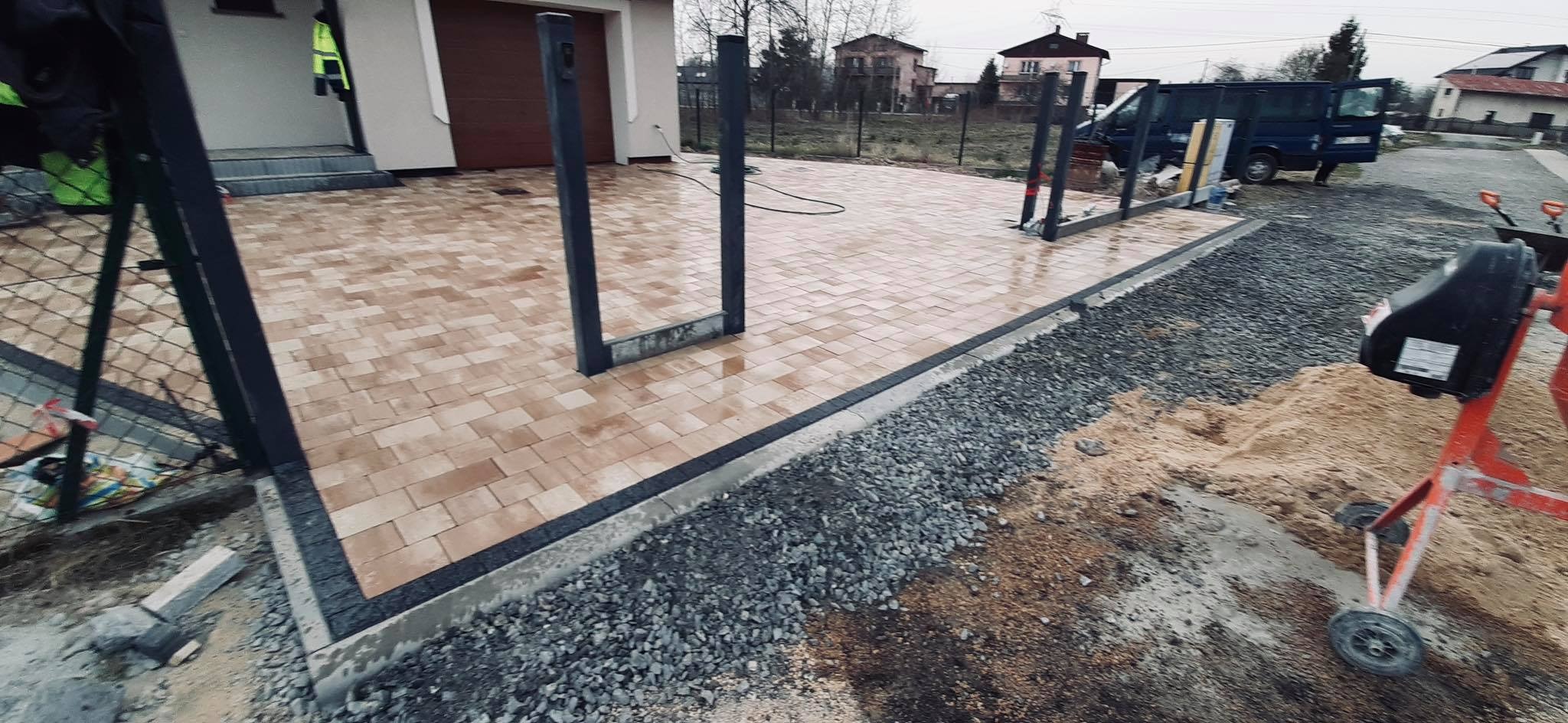 Ułożona beżowa kostka brukowa z czarnym obramowaniem, widoczne przygotowania do montażu ogrodzenia, obok leży rozsypany żwir i stoi mieszarka do betonu, w tle dom jednorodzinny i częściowo widoczny...