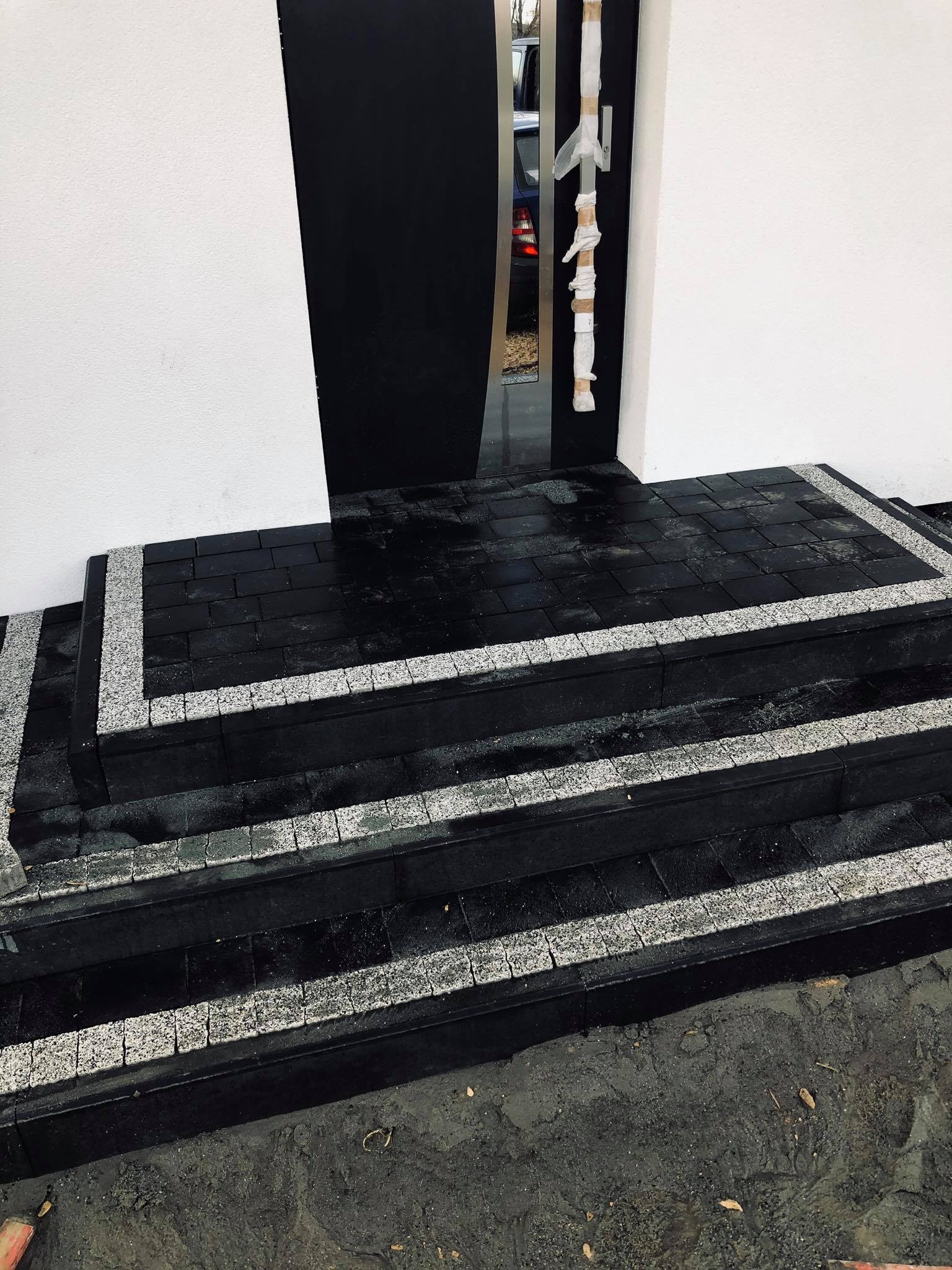 Schody wejściowe do domu wyłożone czarną kostką brukową z obramowaniem z jasnej kostki granitowej, drzwi wejściowe zabezpieczone folią ochronną.