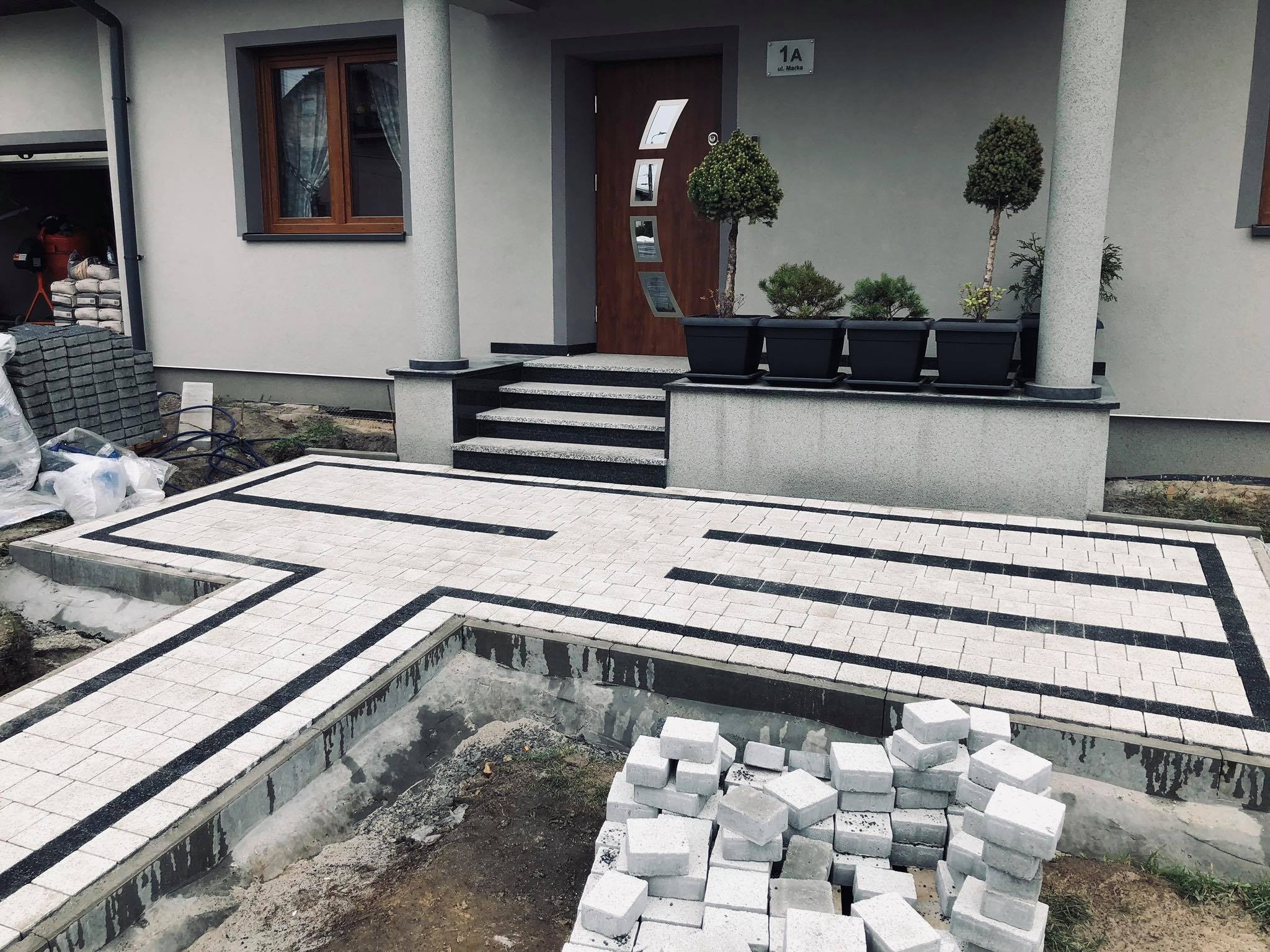 Układanie kostki brukowej przed domem jednorodzinnym, widoczne fragmenty ułożonej nawierzchni z jasnoszarej kostki z czarnymi wstawkami, paleta kostki brukowej obok wykopu, wejście do domu...