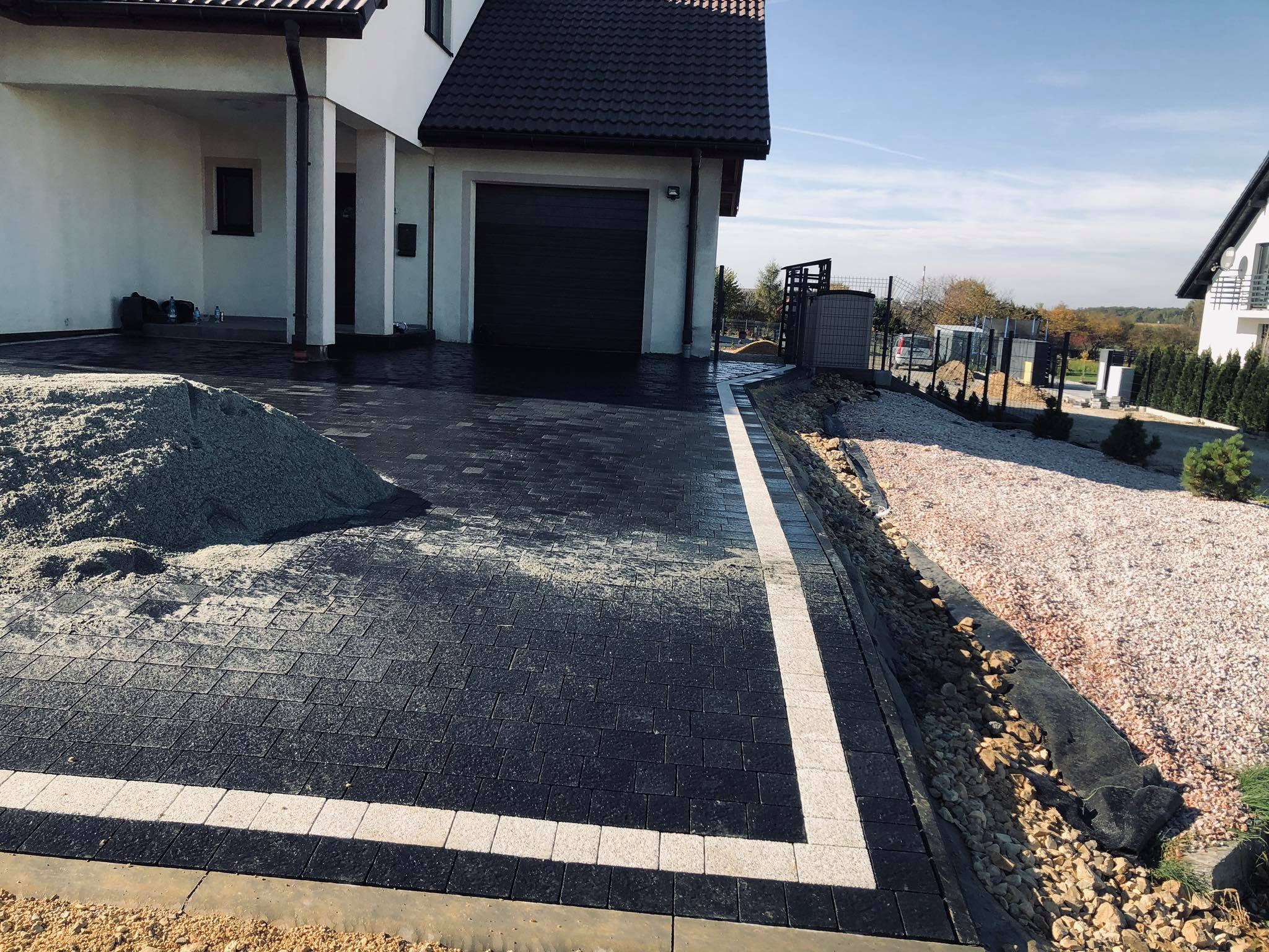 Czarna kostka brukowa z białym obramowaniem przed domem z garażem; widoczny stos piasku i obsypany kamieniami brzeg rabaty.