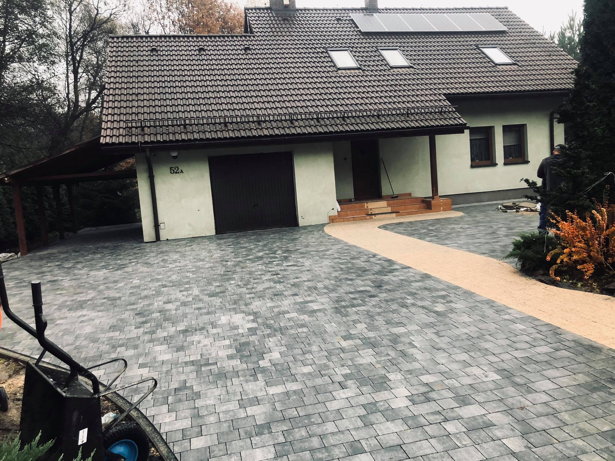 Ułożona szara kostka brukowa na podjeździe przed domem z brązową bramą garażową i panelami słonecznymi na dachu, widoczny fragment taczki i pracownik w tle.