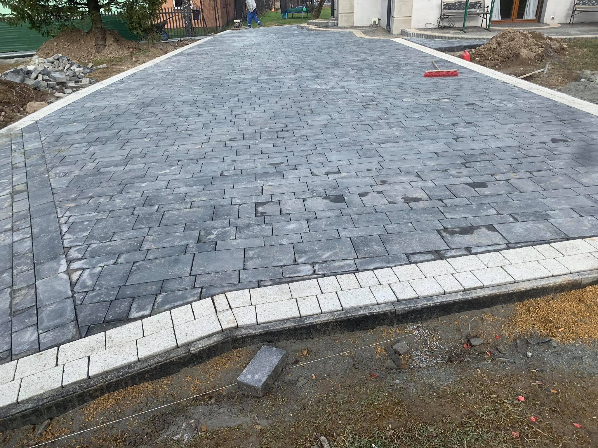 Ułożona nawierzchnia z szarej kostki brukowej z białym obramowaniem, widoczne niedociągnięcia na krawędzi, pojedyncza kostka leżąca obok, oraz czerwona zmiotka w tle.