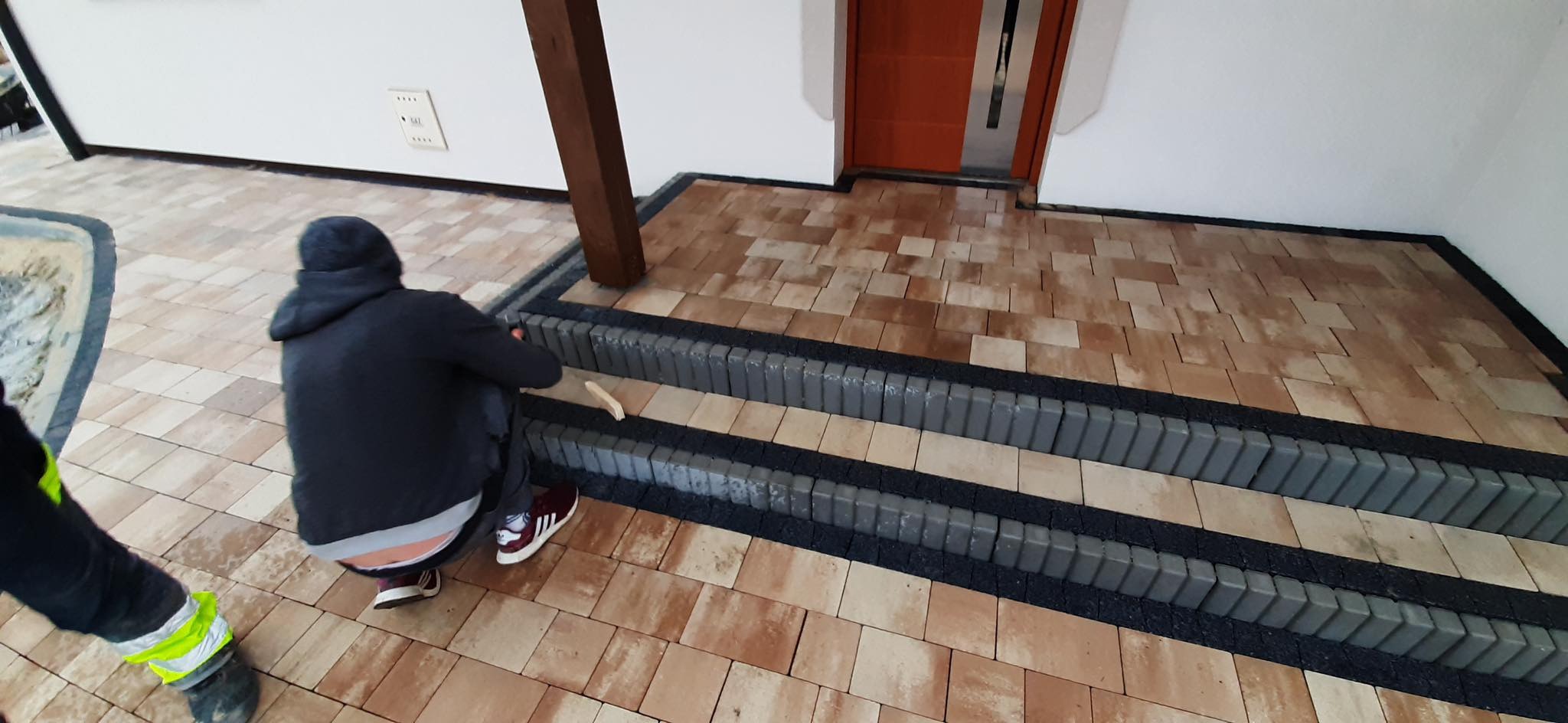 Pracownik w trakcie układania stopni z szarej kostki brukowej na tarasie przed wejściem do budynku, widoczne fragmenty ułożonej już kostki w odcieniach beżu i brązu.