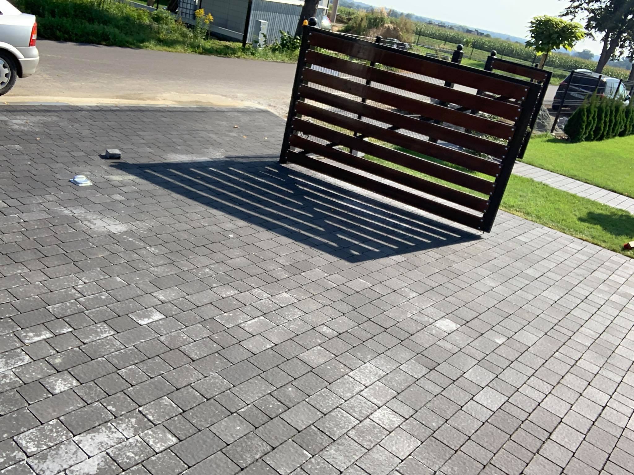 Ułożona szara kostka brukowa na podjeździe z drewnianą bramą w tle, widoczne ślady białego nalotu na kostce.