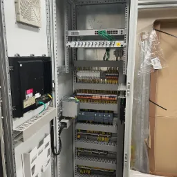 Otwarta szafa sterownicza z widocznym panelem operatorskim Advantech, wentylatorem, listwami zaciskowymi, zasilaczem i elementami automatyki przemysłowej firmy Siemens. Widoczne kable w kolorach...