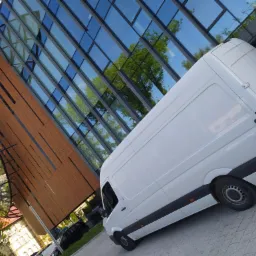 Biały samochód dostawczy marki Mercedes-Benz Sprinter zaparkowany na tle nowoczesnego budynku z przeszkloną fasadą, w której odbijają się drzewa.
