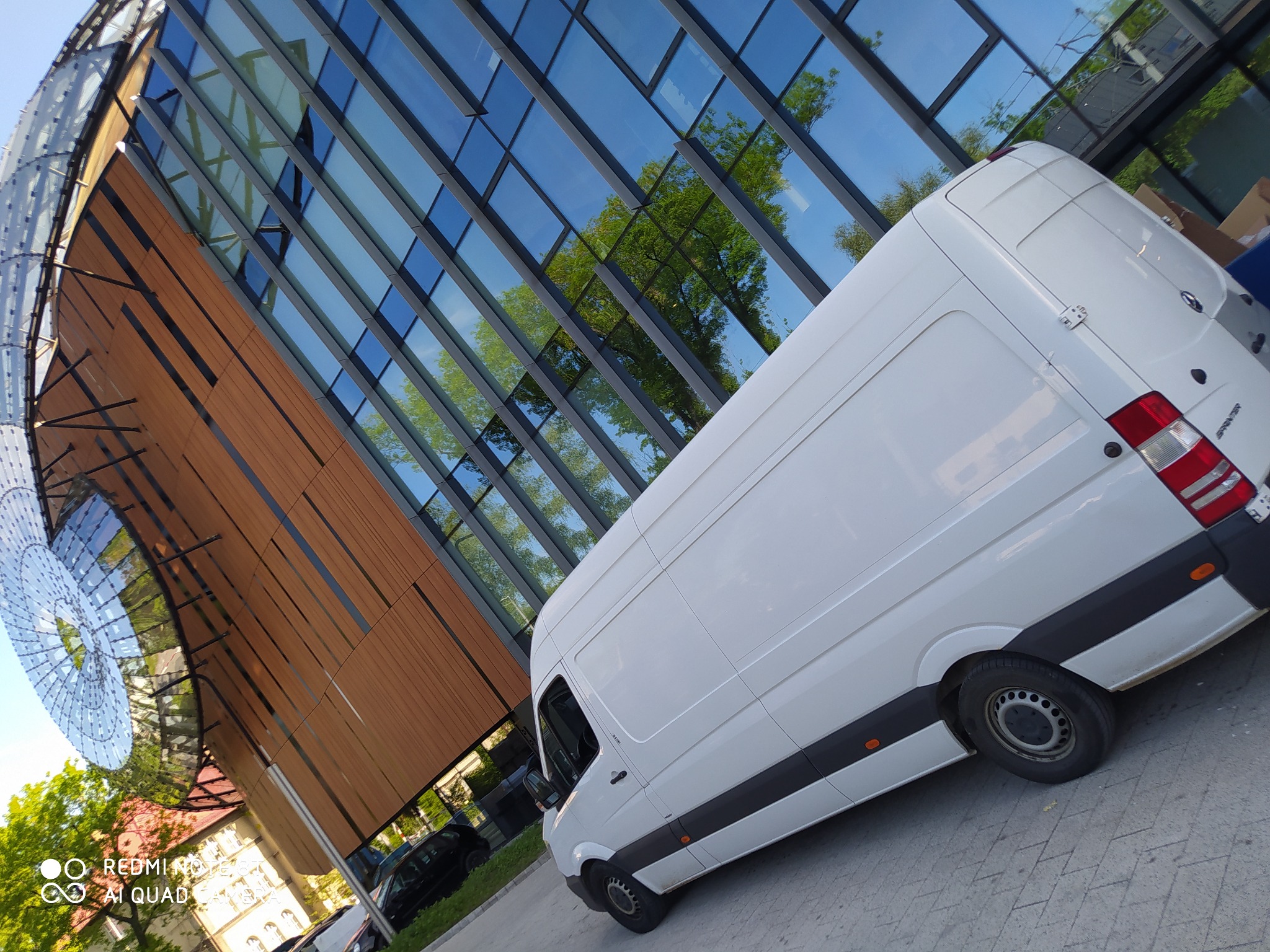 Biały samochód dostawczy marki Mercedes-Benz Sprinter zaparkowany na tle nowoczesnego budynku z przeszkloną fasadą, w której odbijają się drzewa.
