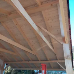 Wnętrze konstrukcji drewnianej z widocznymi belkami stropowymi i deskami, z zamontowanym oświetleniem, częściowo zadaszone, z elementami czerwonej podnośki na dole kadru.