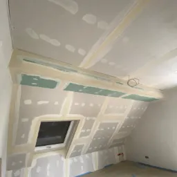 Poddasze w trakcie prac wykończeniowych: płyty gipsowo-kartonowe zamontowane na skosach dachu i ścianach, widoczne łączenia wzmocnione taśmą, okno dachowe, instalacja elektryczna wystająca z sufitu...