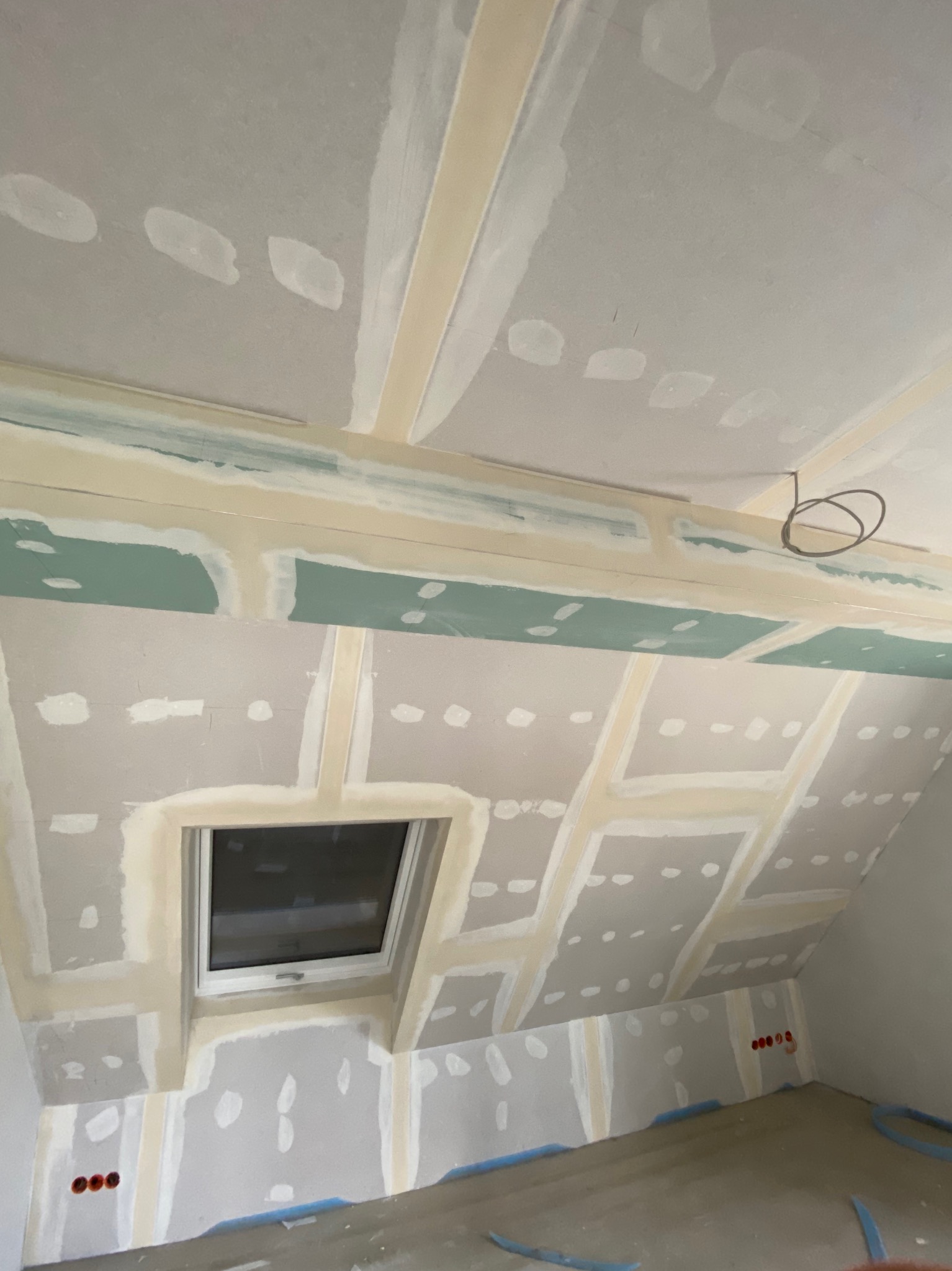 Poddasze z oknem dachowym w trakcie prac wykończeniowych: płyty kartonowo-gipsowe zamontowane, widoczne łączenia wzmocnione taśmą i wstępnie zaszpachlowane, ściany przygotowane do dalszych prac.