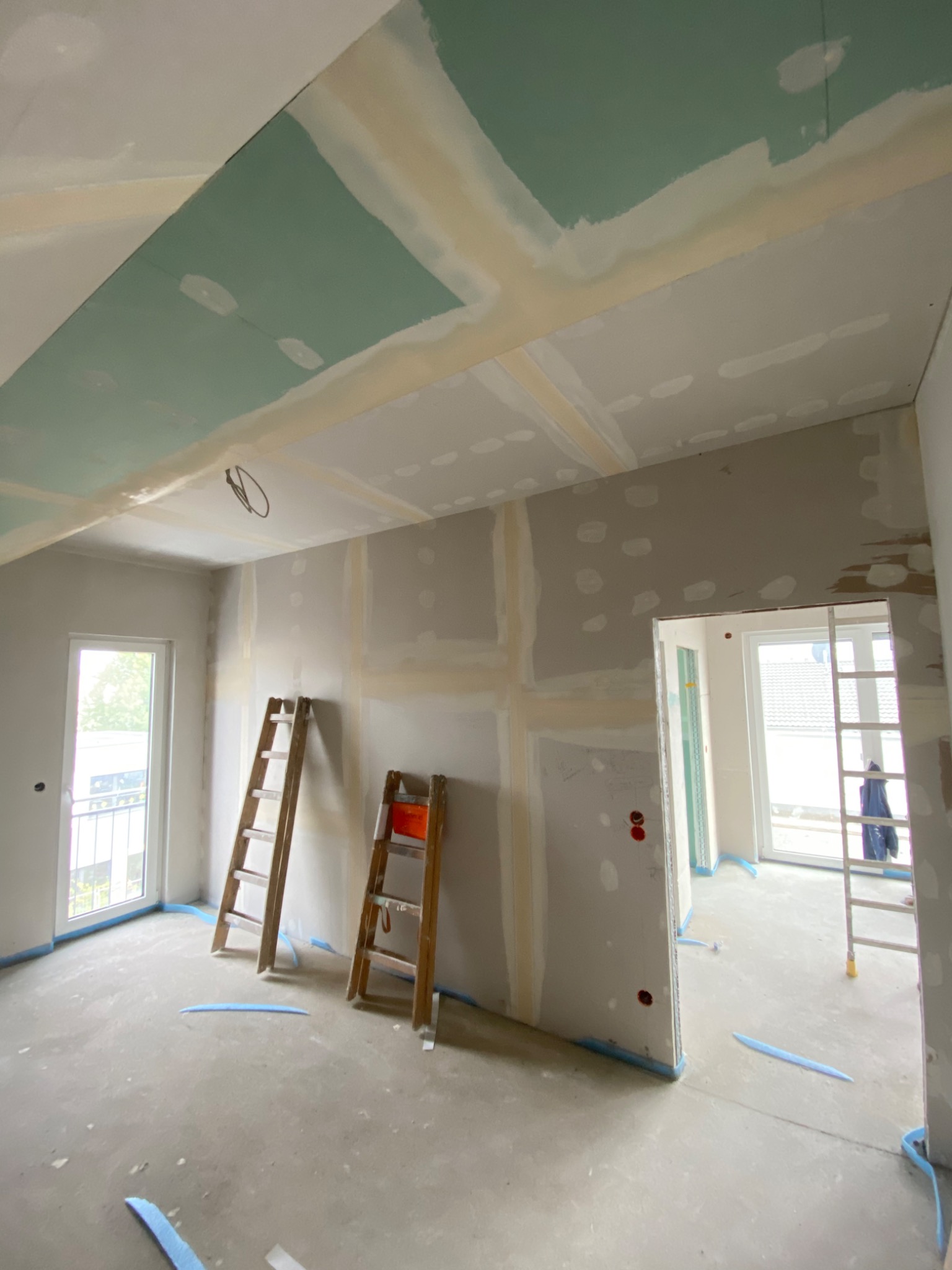 Wnętrze w trakcie remontu: Sufit i ściany z płyt karton gips, częściowo zaszpachlowane, zabezpieczone niebieską taśmą malarską, dwie drewniane drabiny w pomieszczeniu.