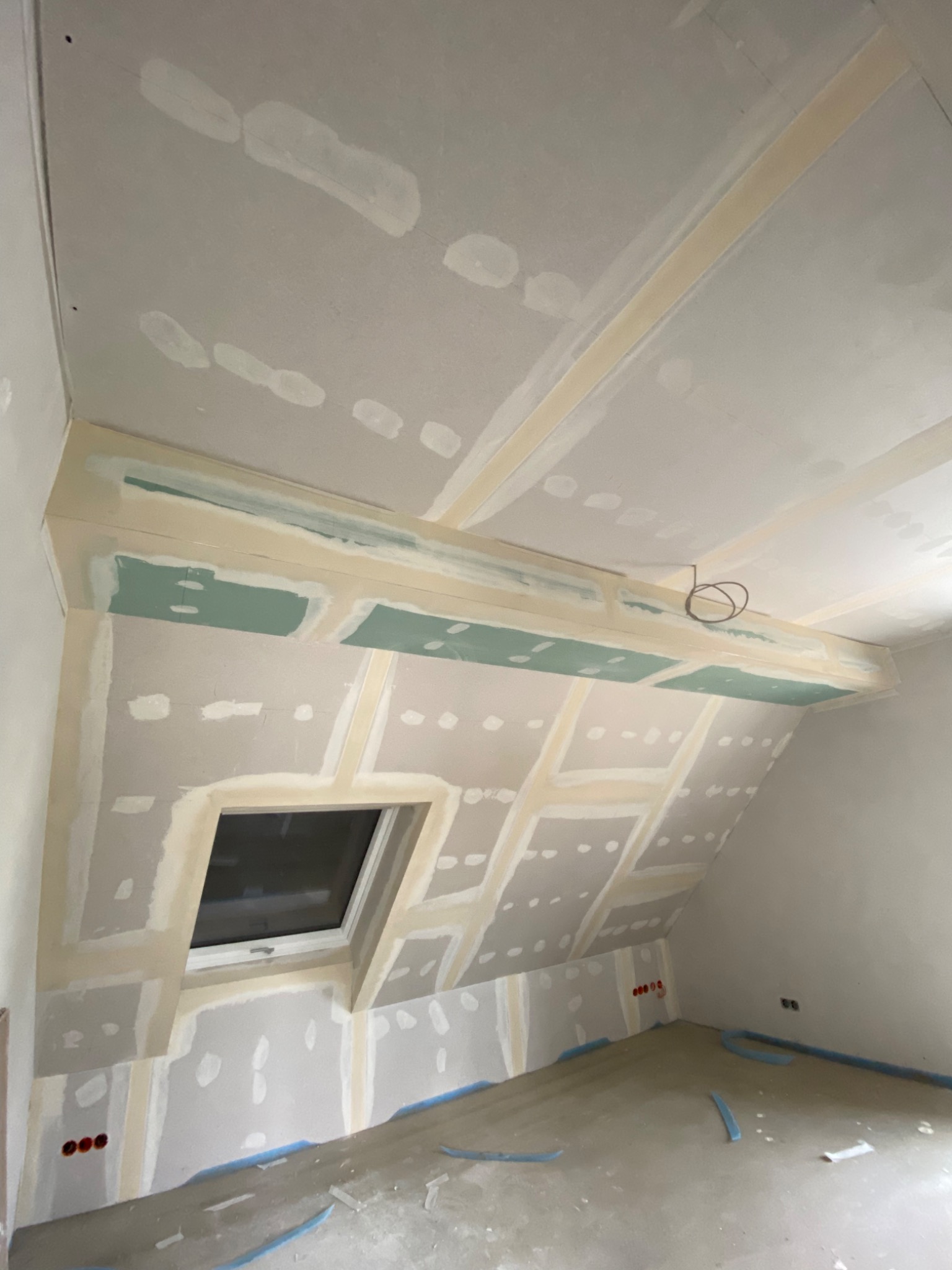 Poddasze w trakcie prac wykończeniowych: płyty gipsowo-kartonowe zamontowane na skosach dachu i ścianach, widoczne łączenia wzmocnione taśmą, okno dachowe, instalacja elektryczna wystająca z sufitu...