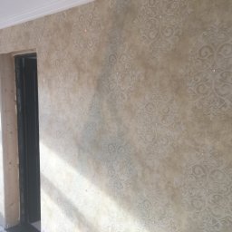 Dawid Fornal - Ściana z dekoracyjną, beżową tapetą w ornamenty, częściowo oświetlona przez promienie słoneczne wpadające przez okno. Widoczny fragment ościeżnicy drzwiowej w trakcie montażu.