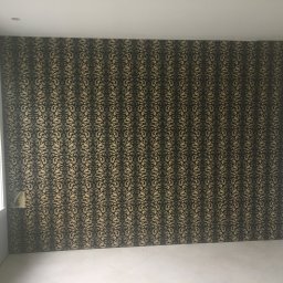 Dawid Fornal - Pokój po położeniu tapety w złoto-czarne ornamenty, widoczny fragment okna i przygotowane narzędzia na parapecie.